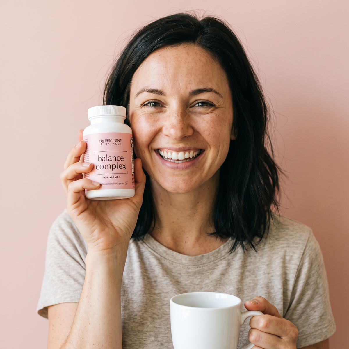 A woman laughing to camera in a heather-oat tee, holding the bottle up beside her face, a white mug in her other hand