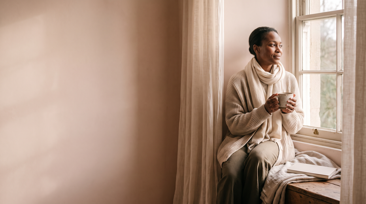 A woman of about 40 on a window seat holding a mug of tea and looking out, a clean blush wall to the left