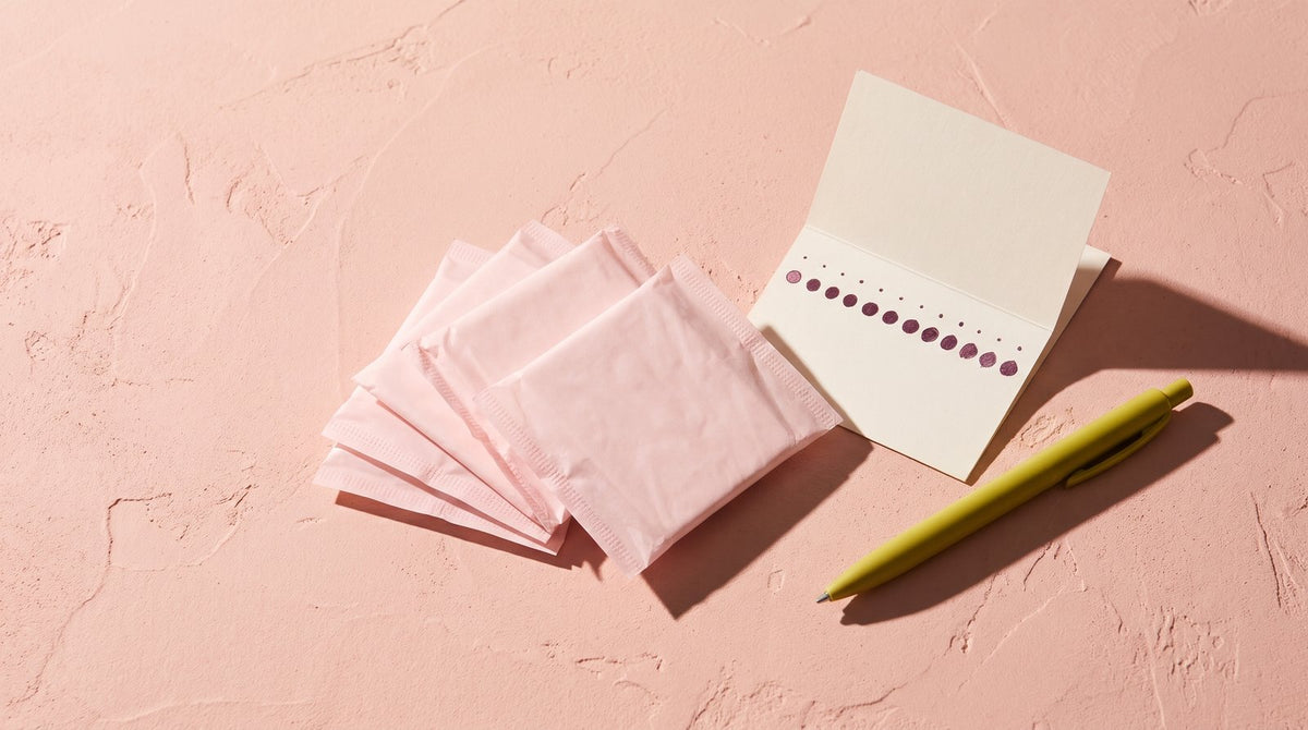 Wrapped pale pink panty liners fanned beside a small cycle card of plum dots and a yellow-green pen on blush plaster