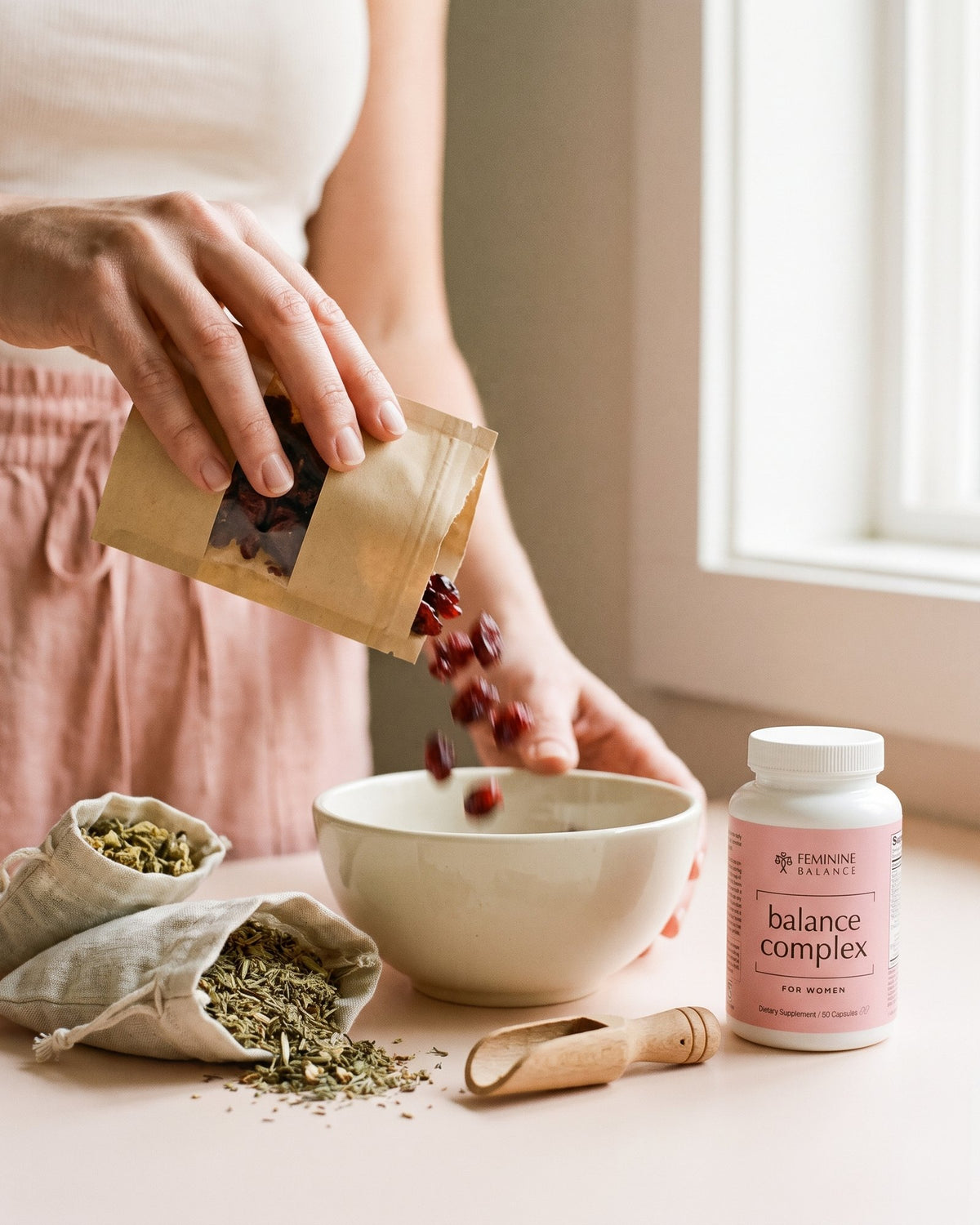 Hands tipping dried cranberries into a bowl beside linen pouches of herbs and the bottle