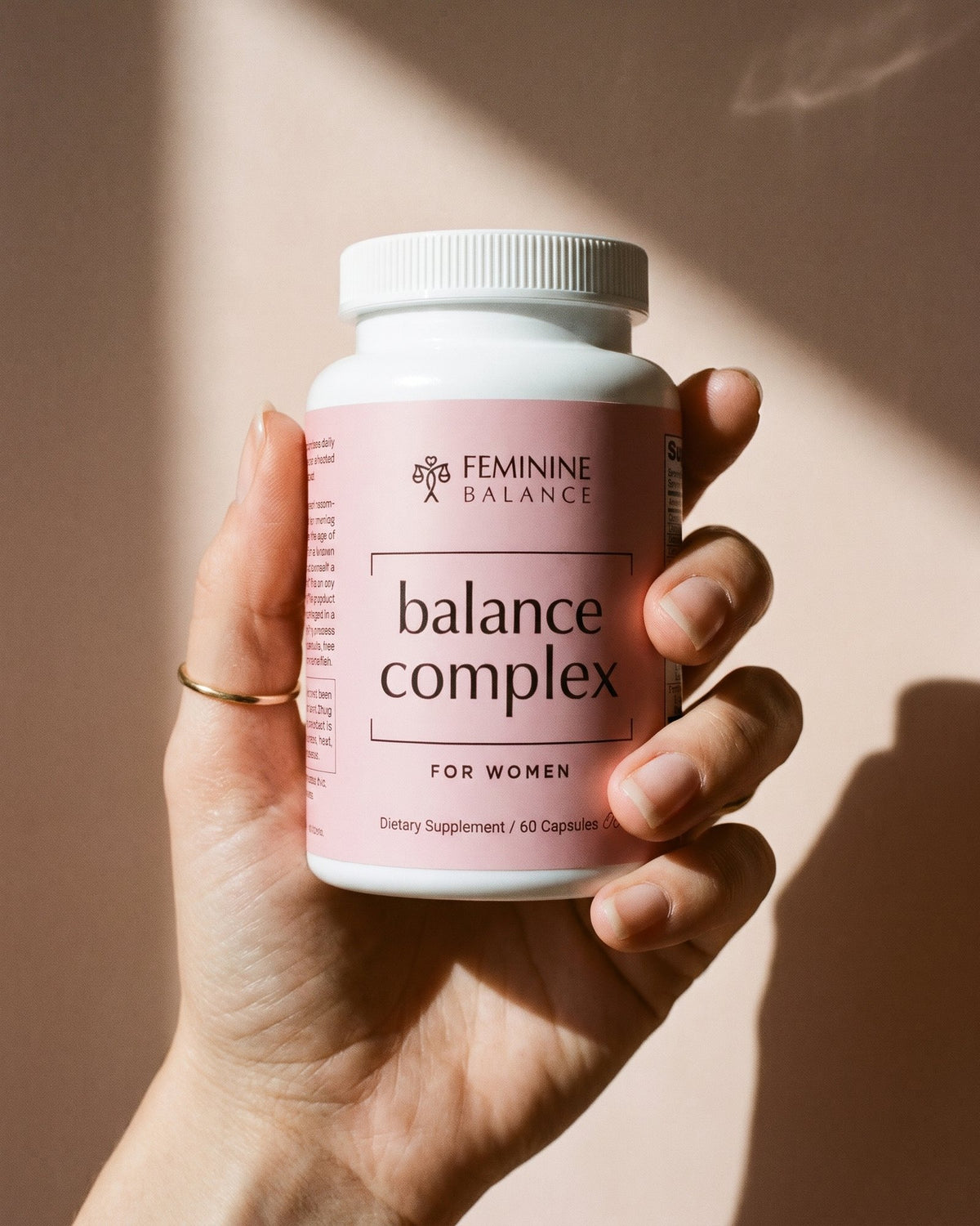 A hand holding the Balance Complex bottle against a plain blush wall in window light