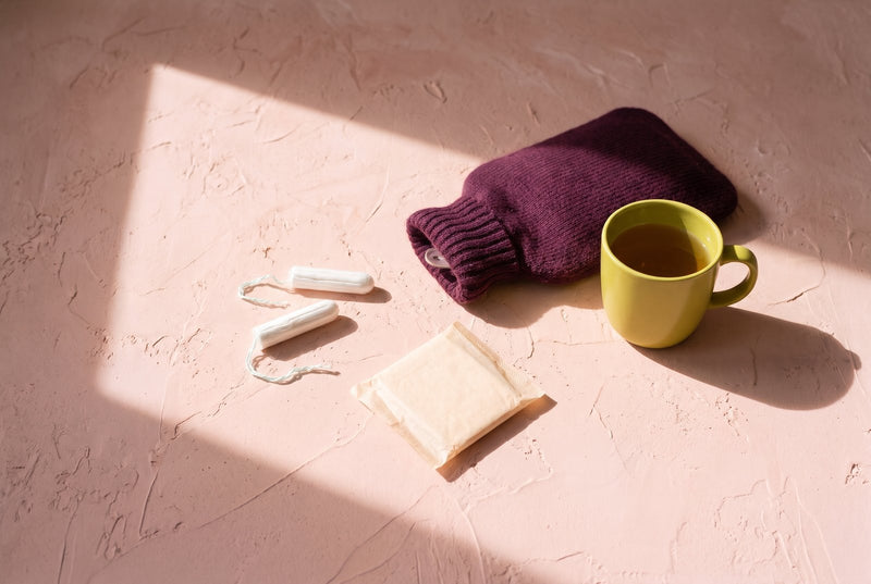Two tampons and a wrapped pad beside a plum knitted hot-water bottle and a yellow-green mug of tea on sunlit blush plaster