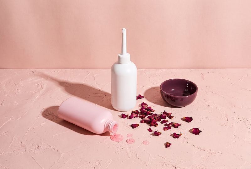A douche bottle, a tipped pink bottle of scented intimate wash with spilled drops, rose petals and a plum bowl on blush plaster