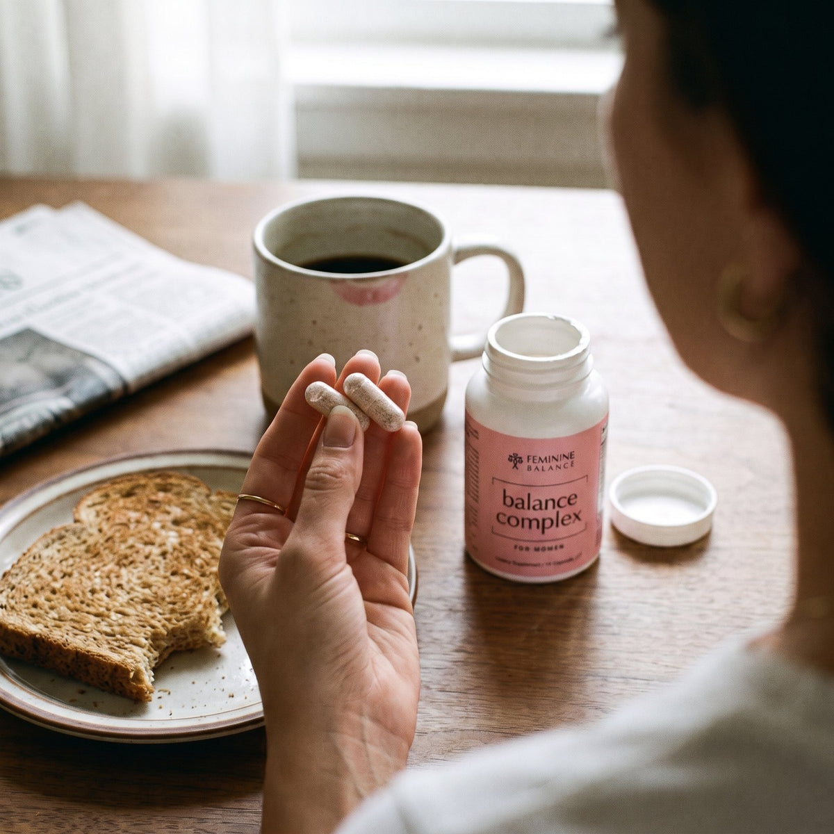 Over-the-shoulder at breakfast: two capsules between her fingers, the open bottle, toast and coffee