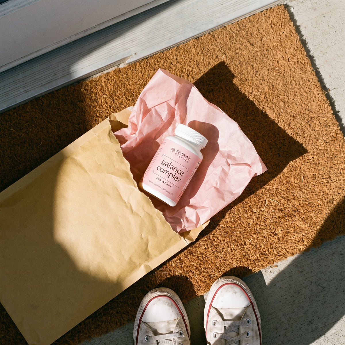 A kraft padded mailer torn open on a doormat, the bottle resting in blush tissue, sneakers at the edge